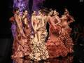Modelos desfilan en el escenario de una pasarela de moda, luciendo vestidos tradicionales flamencos en tonos naranjas y marrones. La iluminación es dramática, enfocándose en los diseños elaborados y las expresiones de las modelos. La audiencia, a oscuras, observa la presentación con interés.