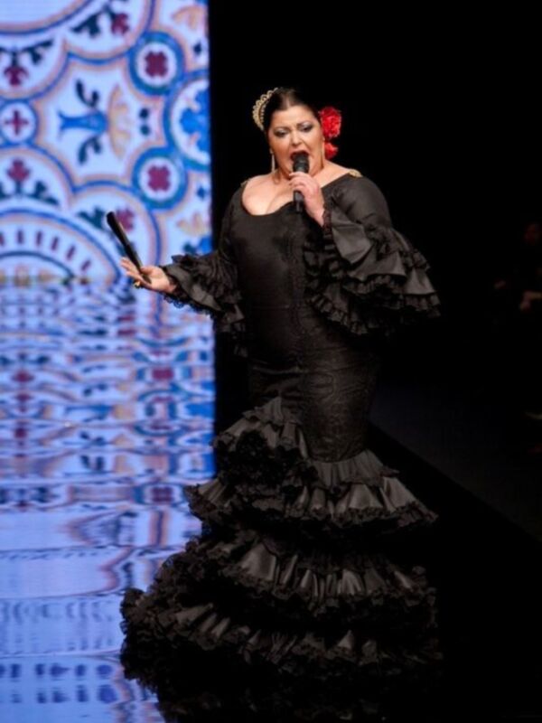 Actriz en traje de flamenca negro con volantes, actuando con micrófono en mano en un escenario con diseño ornamental.