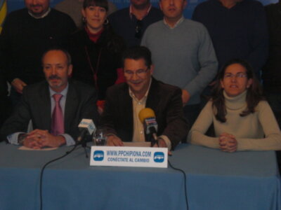 Una reunión formal con varios hombres y una mujer sentados en un escritorio. La imagen muestra a la gente sonriendo o hablando, con algunos hombres vestidos formalmente y una mujer en un suéter blanco. En el escritorio hay un microfono, un papel con texto y una bandera amarilla. La imagen está iluminada de manera uniforme, probablemente en un salón o auditorio.