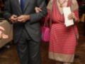 Un hombre en traje negro y corbata azul posa junto a una mujer mayor con vestido rojo a rayas y un bolso rosa. Ambos parecen estar en una reunión formal, posiblemente una celebración o evento.