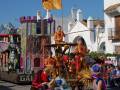 Un carro de carnaval colorido con figuras doradas y un caballo, frente a una casa blanca con tejados de tejas.