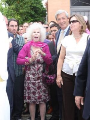 Una anciana con vestido floral y gafas de sol, rodeada por personas en un ambiente formal.