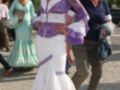 Una mujer vestida con un traje de flamenca blanco y morado, posando en una calle con palmeras al fondo. Detalle de la moda tradicional española.