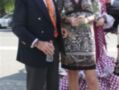 Un hombre y una mujer posan juntos en un evento formal. El hombre lleva un traje negro con corbata naranja y gafas de sol, mientras que la mujer usa un vestido con estampado animal y un sombrero de fiesta. Ambos parecen estar disfrutando del evento.