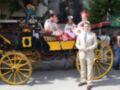 Un hombre en traje beige posa frente a un elegante carruaje amarillo y negro, mientras una mujer en vestido blanco y sombrero de fiesta se sienta detrás. El entorno parece ser una celebración o evento formal, con otros personajes vestidos de manera similar en el fondo.
