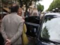 Una pareja se despide de una ceremonia, el hombre le da la mano a la mujer mientras ella sale del coche. Detrás de ellos, se pueden ver personas con sombreros y trajes históricos. La escena parece ser en una calle de un lugar histórico, con árboles y edificios antiguos al fondo.