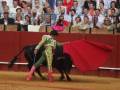 Un torero en el centro de la plaza de toros, con una muleta en la mano y un toro negro detrás de él, mientras el público observa desde las gradas.