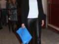 Una mujer con un blazer negro, una camiseta blanca y pantalones de cuero negro. Lleva zapatos negros con tacones altos y una cartera azul. La imagen se toma en un ambiente con paredes de ladrillo y una puerta metálica.