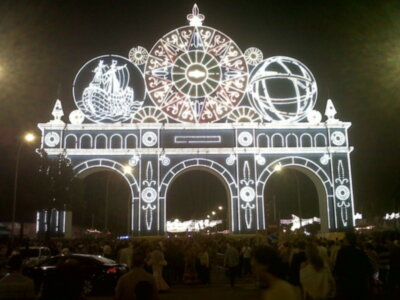 Arco iluminado con motivos navideños y globos, rodeado de una multitud en una feria nocturna.