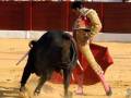 Un torero con capa roja y muletas de color rosa se enfrenta a un toro negro en una plaza de toros. El ambiente es intenso y dramático, con el suelo cubierto de arena y una valla roja en el fondo.