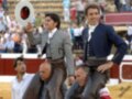 Dos hombres en trajes tradicionales de corrida de toros se preparan para una carrera, mientras que un tercer hombre observa desde el lado. El fondo muestra a espectadores en la gradas del estadio.