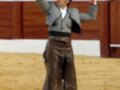 Un torero en traje tradicional, con un sombrero rojo y una chaqueta gris, levanta el capote en la arena de toros. El fondo muestra una pared de madera y a un grupo de espectadores observando la escena.