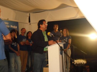 Un hombre en un escenario con una camiseta verde y pantalones azules está hablando al micrófono. A su lado, hay personas en camisetas de color azul y negro escuchando atentamente. La imagen está iluminada por una luz brillante detrás de ellos, y hay un fondo con una bandera azul y blanca.