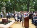 Inauguración de una estatua en un parque con una mujer hablando en un podio y una multitud observando.