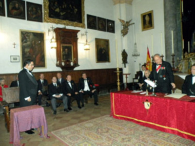 Conferencia o ceremonia formal en un salón decorado con pinturas históricas y bandera española.