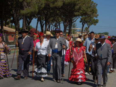 Una procesión tradicional en un ambiente soleado, con personas vestidas de manera distintiva y sombreros típicos. La imagen muestra a varias personas caminando en una calle, algunos con vestidos largos y otros con trajes formales. La escena está ambientada en un día soleado, con árboles y edificios al fondo.