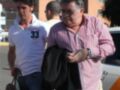 Dos hombres caminan juntos, uno con una chaqueta negra y el otro con un polo blanco. El hombre en la chaqueta parece ser acompañado por el otro, que lleva una mochila. La escena se desarrolla en un entorno urbano con edificios y vehículos al fondo.