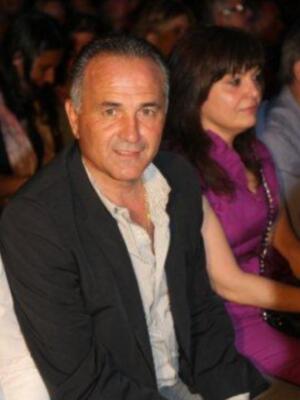 Persona masculina en traje negro y camisa blanca, sentado en un evento público con una audiencia en el fondo.