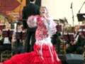 Una mujer en un vestido rojo y blanco se encuentra en el centro de una orquesta, con un hombre detrás de ella. La escena está iluminada y parece ser una presentación musical en un auditorio.