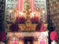Una estatua de la Virgen María en un altar adornado con flores y velas, rodeada por una estructura de madera con patrones geométricos. La escena transmite un ambiente religioso y festivo, probablemente durante una celebración o procesión.