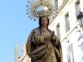 Estatua dorada de una Virgen con un halo y corona, frente a edificios blancos.