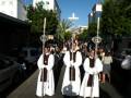Fotografía de una procesión religiosa con tres hombres vestidos con hábitos blancos y negros, portando cruces de plata. El entorno muestra una calle con edificios y coches, bajo un cielo azul claro.
