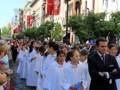 Una procesión religiosa en una calle con edificios históricos. Personas vestidas de blanco y un hombre en traje negro a su lado. Banderas rojas colgadas en los edificios.