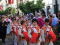 Desfile en una plaza con personas vestidas de trajes tradicionales, banderas y árboles verdes en el fondo.