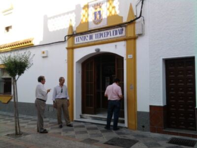 Tres hombres charlan en la entrada de un edificio blanco y amarillo con una escudo en el techo.