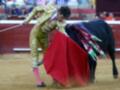 Un torero en traje dorado y rosa se enfrenta a un toro negro en una plaza de toros. El torero sostiene un capote rojo y se prepara para el combate.