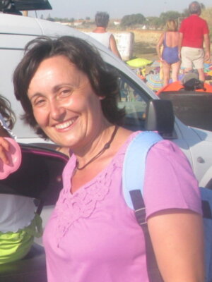 A una mujer sonriendo mientras se sienta en el asiento de atrás de un vehículo. Está vestida con una camiseta púrpura y tiene un bolso azul sobre los hombros. En el fondo, se pueden ver personas y una playa con sombrillas y playas de arena.