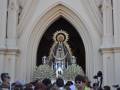 Una procesión religiosa con una estatua de la Virgen María en el centro, rodeada por flores blancas y una multitud de personas observando la ceremonia. La arquitectura del edificio en el fondo es gótica, con una gran entrada de arco. La escena transmite un ambiente festivo y religioso.