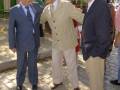 Tres hombres en trajes de negocios se encuentran en una calle con un fondo verde y edificios. Uno de ellos sostiene la mano del otro, mientras que el tercero observa desde atrás. La escena parece ser formal y posiblemente de una reunión o evento empresarial.