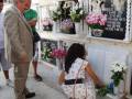 Una persona en un traje beige se inclina hacia una tumba con flores y un letrero negro. La tumba tiene nombres y fechas en letras blancas. Algunos individuos observan la escena, uno de ellos con una mochila y otro con un bolso. La tumba está decorada con flores en macetas de diferentes tamaños y colores.