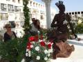 Una escultura de un hombre negro sentado, rodeada por flores y una mujer en el fondo.