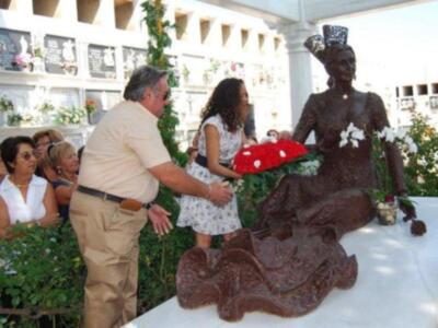 Una figura de chocolate con forma de una mujer sentada, vestida con un vestido negro y sombrero de copa, está en una tumba. Algunas personas están alrededor, incluyendo a un hombre que parece estar tocando la figura de chocolate. El entorno es un cementerio con flores y lápidas en el fondo.