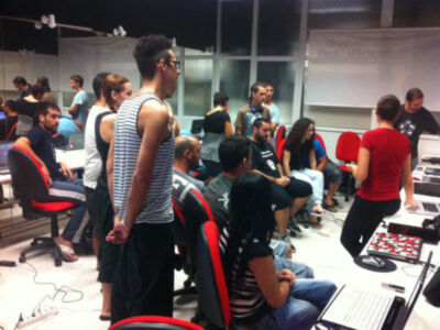 Un grupo de personas en una sala de reuniones colaborando en una actividad de trabajo o discusión, rodeados de tecnología y equipamiento de oficina.