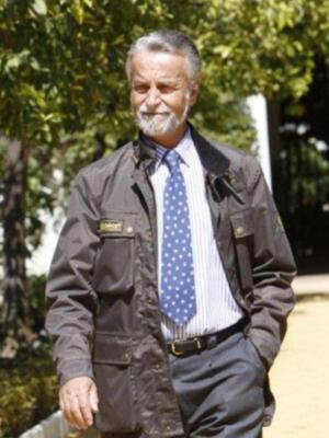 Un hombre con barba y bigote, vistiendo una chaqueta verde militar, corbata azul con patrones y camisa de rayas. Está en un entorno natural con árboles y una valla al fondo.