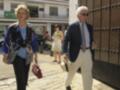 Una pareja de ancianos camina juntos por una calle adoquinada, rodeados de edificios de estilo clásico.