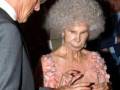 Una anciana con una peluca voluminosa y un vestido rosa es el centro de atención mientras se tocan las manos con otro hombre. La escena parece ser una celebración o evento formal, con un fondo que incluye azulejos y una silla.