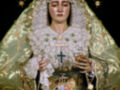 Imagen de una Virgen con un vestido verde y dorado, coronada con una corona de oro. La Virgen tiene un manto blanco y lleva en sus manos un símbolo religioso.