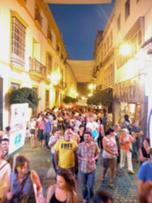 Una multitud camina por una calle de noche iluminada, con edificios históricos a ambos lados. La gente parece disfrutar de una celebración o evento, con algunos portando pancartas y camisetas. La atmósfera es festiva, con luces brillantes y un cielo nocturno claro.