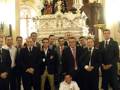 Un grupo de hombres en trajes posa frente a una imagen religiosa en un interior iluminado.