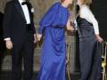Una mujer en un vestido azul sosteniendo la mano de otra mujer, ambas sonríen. Detrás de ellas, un hombre en traje negro con corbata y camisa blanca. La escena parece ser una celebración formal en un entorno interior con luces y decoraciones.