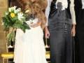 Dos mujeres posan juntas, una con un vestido blanco y la otra con un vestido negro, ambos con accesorios elegantes. Una de las mujeres sostiene un ramo de flores. Detrás de ellas, se puede ver una silla con un asiento amarillo y un fondo oscuro.