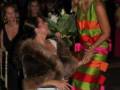 Una mujer en un vestido blanco y una peluda chaqueta marrón se inclina hacia abajo para recibir flores de otra mujer vestida con un vestido de colores vibrantes. Ambas parecen estar en una ceremonia formal, posiblemente un evento de premios.