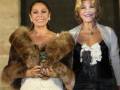 Dos mujeres posan juntas en un evento formal, una con un abrigo de piel marrón y la otra con un vestido negro.