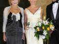 Una mujer en un vestido negro con una corbata blanca, otra en un vestido blanco con un ramo de flores y un hombre en traje negro.