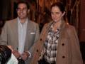 Un hombre y una mujer caminan juntos por una calle iluminada. El hombre lleva un traje claro y la mujer una chaqueta beige con una camisa a cuadros. La imagen sugiere un ambiente nocturno urbano.