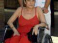 Una mujer en una silla de ruedas, vestida con un vestido rojo, posa para la foto. Detrás de ella, una persona con un uniforme médico sostiene su brazo.
